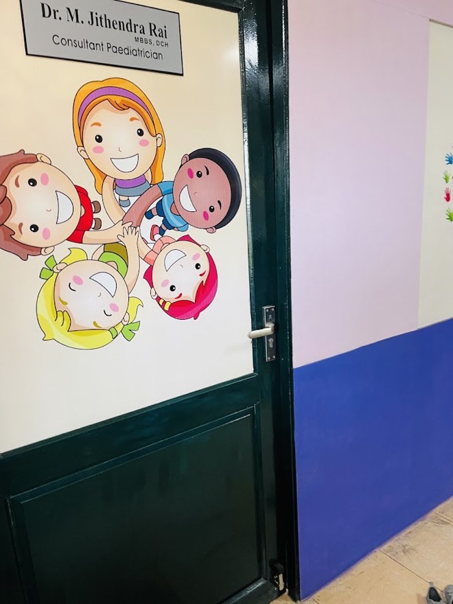 The consulting room door at KINDER clinic, decorated with a cheerful illustration of smiling children and the nameplate of Dr. M. Jithendra Rai, Consultant Paediatrician.